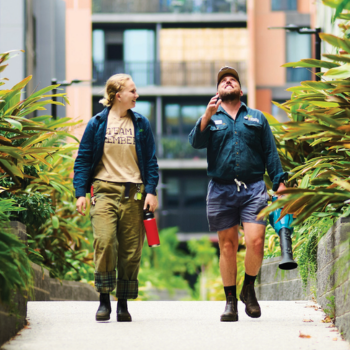 Two people walking and chatting along a lush urban garden path between apartment buildings on a sunny day